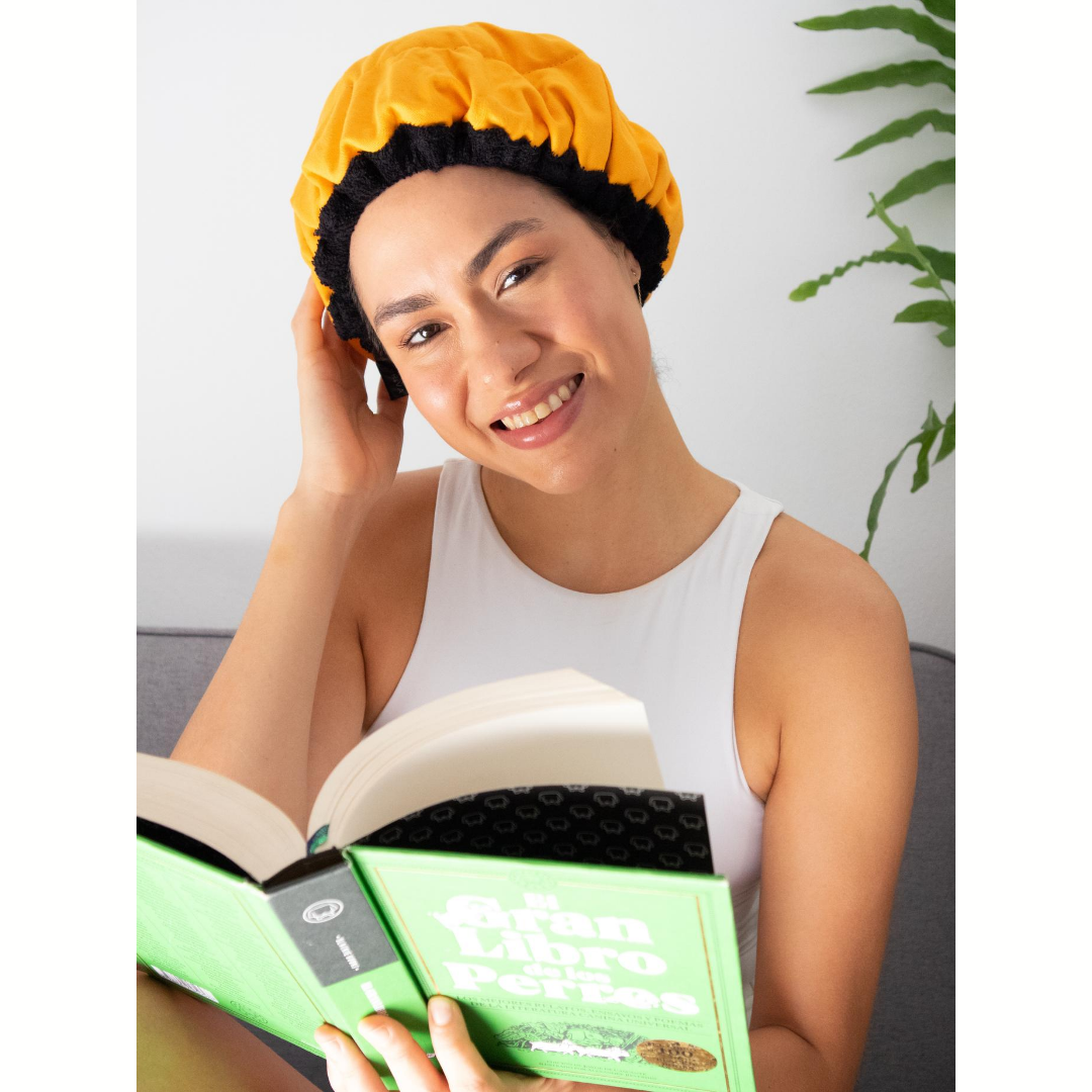 Woman wearing yellow microwavable flaxseed-filled deep conditioning heat cap while reading book and smiling